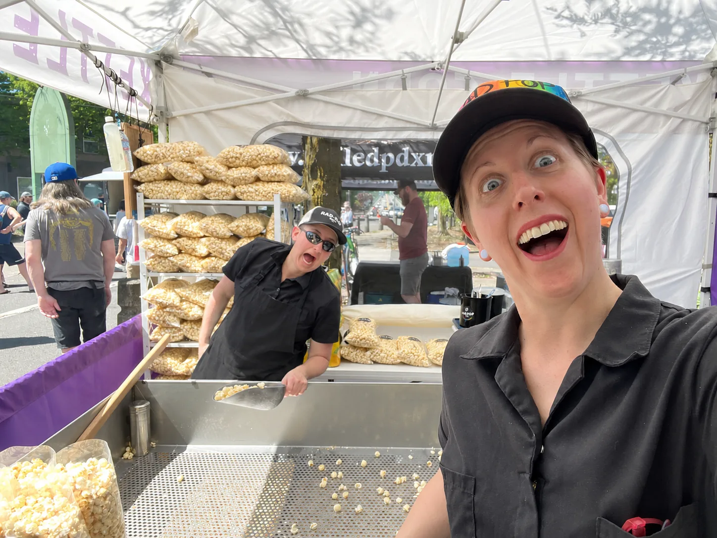 Katie and Iggy smiling inside the Rad Pop market booth with shelves full of popcorn bags